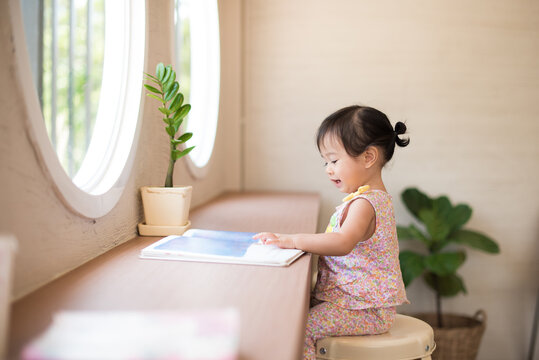 Cute little child girl reading a book in the room near window.