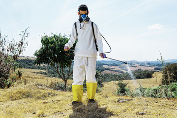 Latin man wearing appropriate clothing and protective mask spraying weeds in terrain