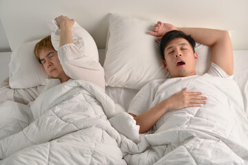  Asian man snoring loudly while his wife is covering ears with the pillow