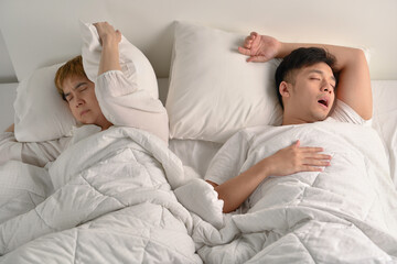 Asian man snoring loudly while his wife is covering ears with the pillow
