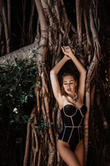 Young slim woman in black transparent bodysuit, swimsuit and jewelry stands leaning at branches of tropical tree, lianas sexy posing holding hands up during vacation. Savage. Summer vibes