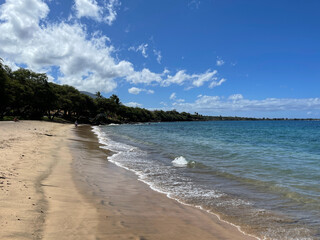 Large puffy clouds hang in a pristine blue sky as ocean waves lap the golden shores of Ka'anapali...