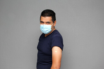 Healthy Latino adult man with mask showing his arm that recently received the Covid-19 vaccine
