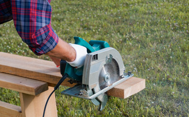 Working with a power tool. A builder is sawing a board at the construction site of a new house. Close-up on gloved hands holding a circular saw. - obrazy, fototapety, plakaty