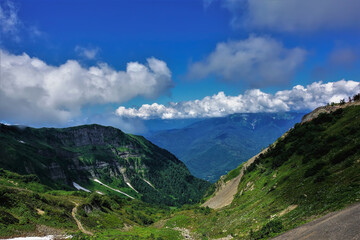 Fototapeta na wymiar Green grass and wildflowers grow on the mountain slopes. Patches of melted snow are visible among the greenery. There are picturesque cumulus clouds in the blue sky. A sunny summer day. Caucasus