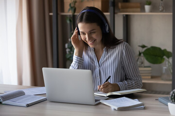 Smiling millennial Caucasian woman in headphones sit on table at home study distant on computer....