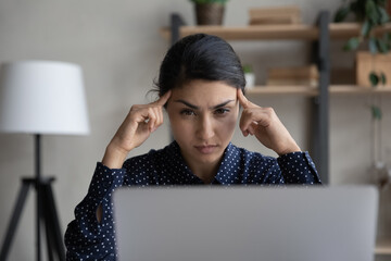 Pensive focused millennial Indian woman look at laptop screen think ponder of problem solution. Concentrated young biracial female employee work on computer make decision or plan at workplace.