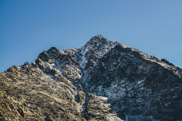 Awesome mountain landscape with black peaked top with white snow in golden sunshine under blue sky. Beautiful snow-covered pointy peak in gold sunlight. Atmospheric mountain scenery with peaked top.