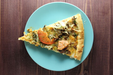 A slice of pizza with tomatoes, herbs and fish on a blue plate and a wooden background. a photo for the menu or a poster. top view.