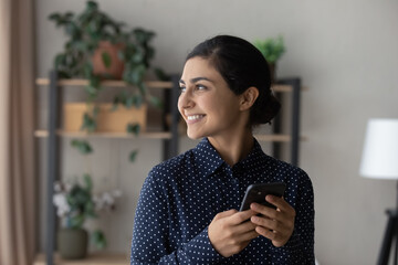 Smiling millennial Indian woman use smartphone look in window distance dreaming or thinking. Happy young biracial female speak on video webcam call on cellphone make plans or visualize.