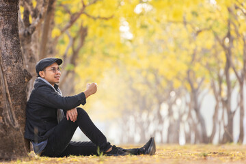 Asian man holding cup of coffee while sitting outdoor under the ginkgo tree at the public park during autumn season while the leaf turning into yellow