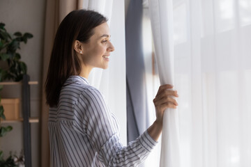 Happy millennial Caucasian woman open curtains look in window meet new bright day at home. Smiling young female renter think dream or future perspective opportunities. Visualization concept.