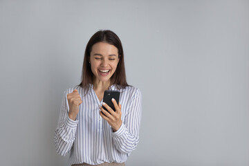 Smiling young Caucasian woman isolated on grey studio background triumph reading good news on smartphone. Happy millennial female feel euphoric excited with lottery won victory on cellphone.