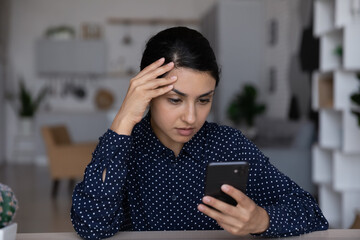 Pensive unhappy millennial Indian woman look at cellphone screen think of web problem or issue on...