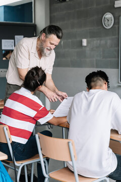 Senior Teacher with beard helping student. Friendly Professor explaining exam exercise to teenager students in college high school classroom