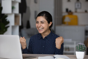 Overjoyed young Indian woman sit at desk at home office feel euphoric win online lottery on laptop. Smiling millennial biracial female look at computer screen triumph reading good news on web.