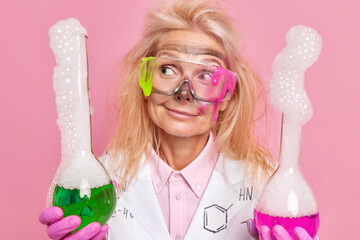 Thoughtful woman biochemist wears white coat protective glasses holds two flasks with explosed...