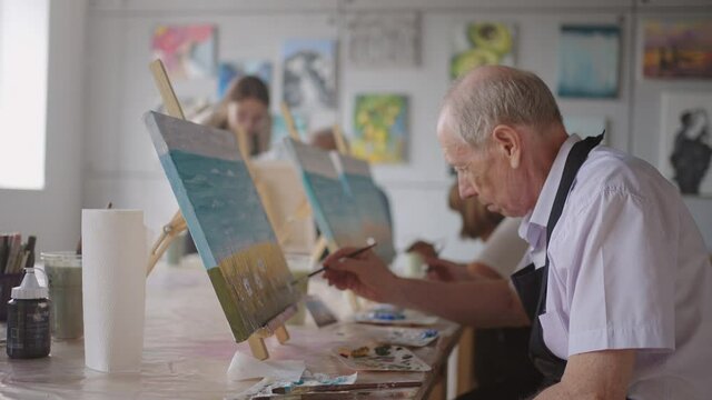 A young woman teacher helps an old pensioner learn to paint a picture. Senior man learning to draw