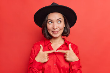 Pensive shy woman feels indecisive before asking risky question gestures and looks away wears stylish black hat and shirt poses against vivid red background. Asian female makes is for me gesture