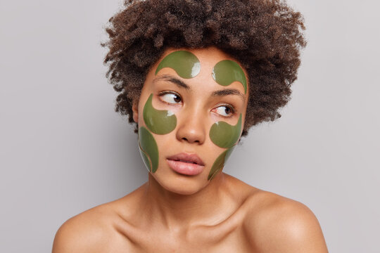 Photo of serious curly haired young woman with pensive expression applies hydrogel green patches on face to remove wrinkles and fine lines stands bare shoulders isolated over grey background