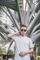 Portrait of handsome young man standing in the park near palm branches. Wearing white t-shirt and sunglasses. Mock-up white t-shirt. High quality