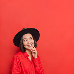 Vertical shot of thoughtful young Asian woman has glad face expression keeps hand on face focused above dressed in fashionable outfit poses against vivid red background copy space for your promo