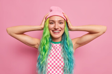Obraz na płótnie Canvas Positive European teenage girl keeps hands to head happy to buy new outfit has dyes colorful hair has piercing in nose isolated over pink background. Glad stylish woman shows her trendy hairstyle