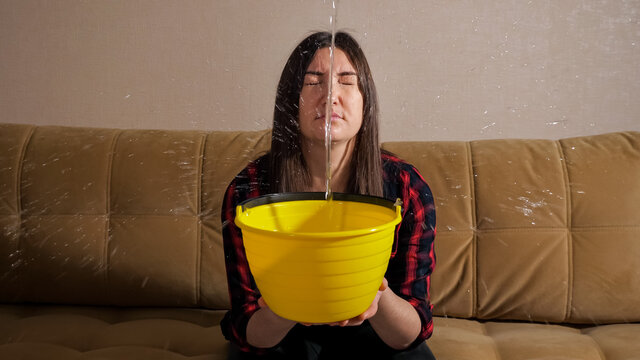 Shocked Brunette Woman In Checkered Shirt Collects Water Flowing From Upstairs Neighbors Into Yellow Bucket On Sofa In Modern Living Room