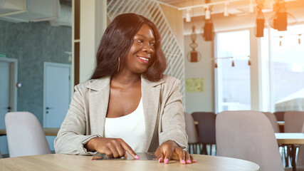 Positive young African-American lady with menu waves hand to call waiter and make order sitting at...