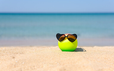 A colored ball with wearing heart-shaped sunglasses lies on a sandy beach against the backdrop of the sea.