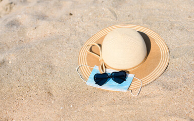 A straw hat with a medical mask and heart-shaped sunglasses lie on the sand. View from above.