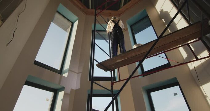 Painter primes the wall, stands high on the scaffolding inside the house. Low angle shot