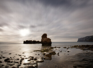 A long exposure on the rocky coast of Denia on the Mediterranean in summer. The water looks smooth and the sun can be seen behind the clouds. In the background the cliffs protrude into the sea.