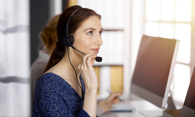 Asian or Latin american businesswoman talking by headset in sunny office. Call center and diverse people group in business