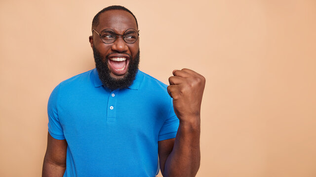 Joyful dark skinned man does winner gesture celebrates victory makes fst bump exclaims from happiness wears round spectacles basic blue t shirt isolated over beige background blank copy space