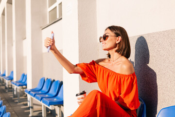 Stylish woman in orange clothes at sunset at cycle track stadium with cup of coffee and mobile phone take photo selfie - obrazy, fototapety, plakaty
