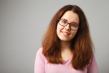 Redhead long-haired girl with glasses smiles, looking at the camera. Studio girl portrait on a solid light background