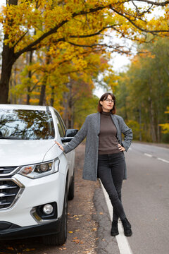 Stylish pretty caucasian woman in glasses, grey coat, brown  sweater and black jeans stands leaning on the SUV car. Autumn forest with yellow leaves on background. Travelling on car, freedom concept. 