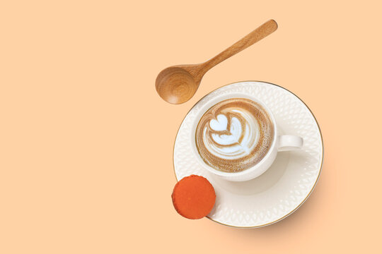 A wooden spoon levitating on a white porcelain cup with cappuccino coffee and a gold rimmed saucer with an orange macaroon.
