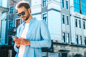 Handsome smiling stylish hipster lambersexual model.Modern man dressed in blue shirt. Fashion male posing on the street background in sunglasses. Outdoors at sunset. Using smartphone apps.Writes sms