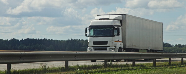 One European white semi trucks front side view into oncoming traffic drive behind the road fence on suburban highway road at summer day on green forest background, cargo transportation logistics