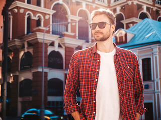 Handsome smiling stylish hipster lambersexual model.Modern man dressed in red checkered shirt. Fashion male posing near skyscraper on the street background in sunglasses. Outdoors at sunset