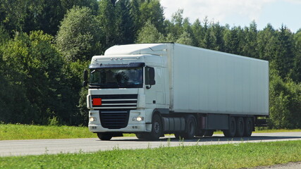 One European white semi trucks front side view into oncoming traffic drive on the suburban highway road at Sunny summer day on green forest background, cargo transportation logistics traffic landscape