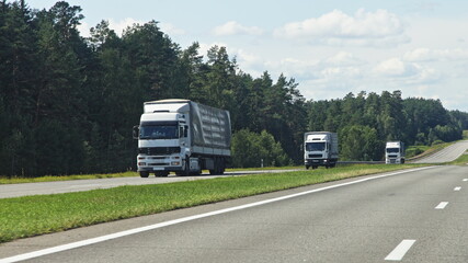 European semi trucks front side view into oncoming traffic drive on the suburban highway road at Sunny summer day on green forest and blue cloudy sky background, traffic landscape