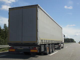 European heavy truck move on the countryside highway road at Sunny summer day on green forest, road fence and blue sky with clouds background, back side view, transportation logistics