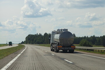 Fototapeta na wymiar Gray barrel 3 axle tanker semi truck back side view on right lane move on the suburban highway road at Sunny summer day on blue cloudy sky background , liquid transportation cargo logistics in Russia