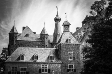 Black and white photo of medieval dutch mystical spooky fairy castle with towers and clock from 14th century, rees against grey sky - Kasteel Doorwerth, Netherlands