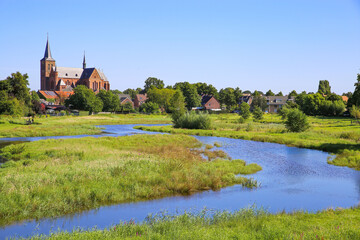 Obraz premium View over green wetland water on cityscape of durch town with tower of medieval church against blue summer sky - Neer (Limburg), Netherlands