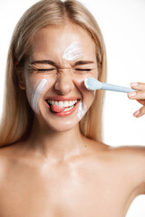 Vertical shot of blond happy girl laughing while using face mask, apply cream or facial lotion,...