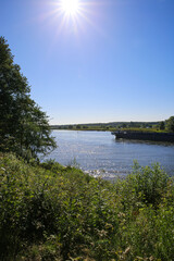 Fototapeta premium View over green rural landscape on river Maas with inland waterway vessel against blue summer sky with sun star - Between Roermond and Venlo, Netherlands